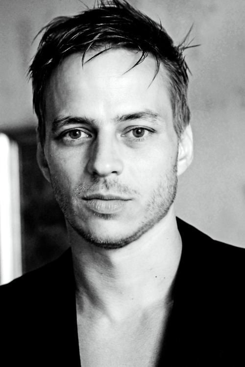 Tom Wlaschiha is serious, has black hair, a beard, and a mustache, with moles on his neck, wearing a black suit.