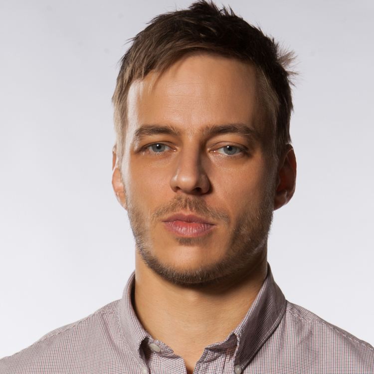 Tom Wlaschiha is serious, has brown hair, a beard, and a mustache, wearing a light-colored checkered polo.