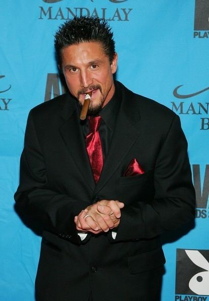 Tommy Gunn with a cigarette in his mouth and wearing a black formal suit along with a red tie.