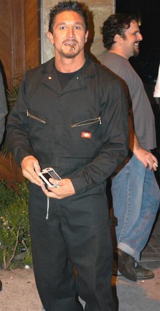 Tommy Gunn with a cigarette in his mouth and holding his phone while wearing a black jacket.
