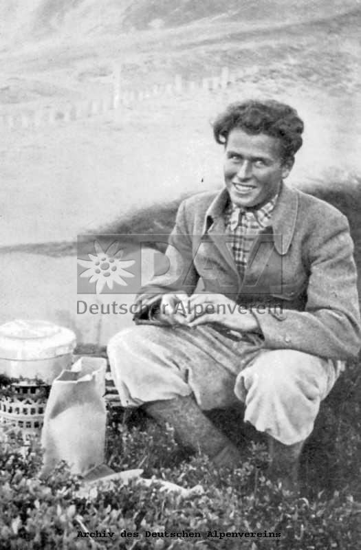 Toni Kurz smiling while sitting on the rock and wearing a coat, checkered long sleeves, pants, high socks, and shoes