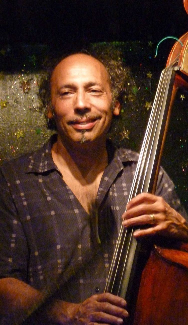 Tony Garnier smiling while holding a guitar, with curly hair, and wearing a checkered gray polo shirt.