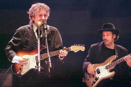 Tony Garnier sings and plays guitar while Bob Dylan plays guitar. Tony wearing a black coat over white long sleeves and a black tie while Bob wearing a black hat, and a black coat over a black shirt.