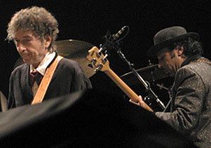 Tony Garnier with a serious face while Bob Dylan plays guitar. Tony wearing a black coat over white long sleeves and a black tie while Bob wearing a black hat, and a black coat.
