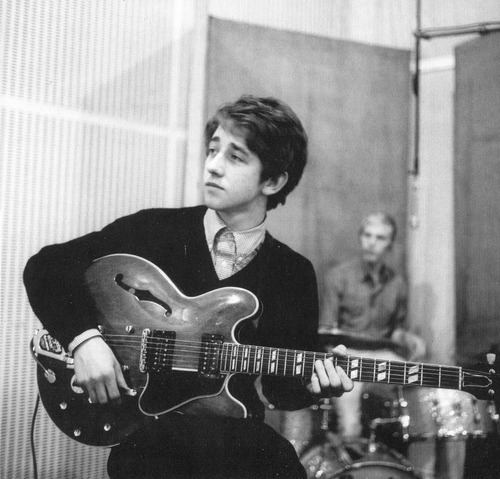 Tony Hicks looking at the left side while playing guitar, behind him is a man playing drums and wearing a long sleeve shirt. Tony is wearing a long sleeve under a black sweater