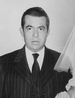 Tony Martinez with a serious face while wearing a white long sleeve under a black necktie and black striped coat