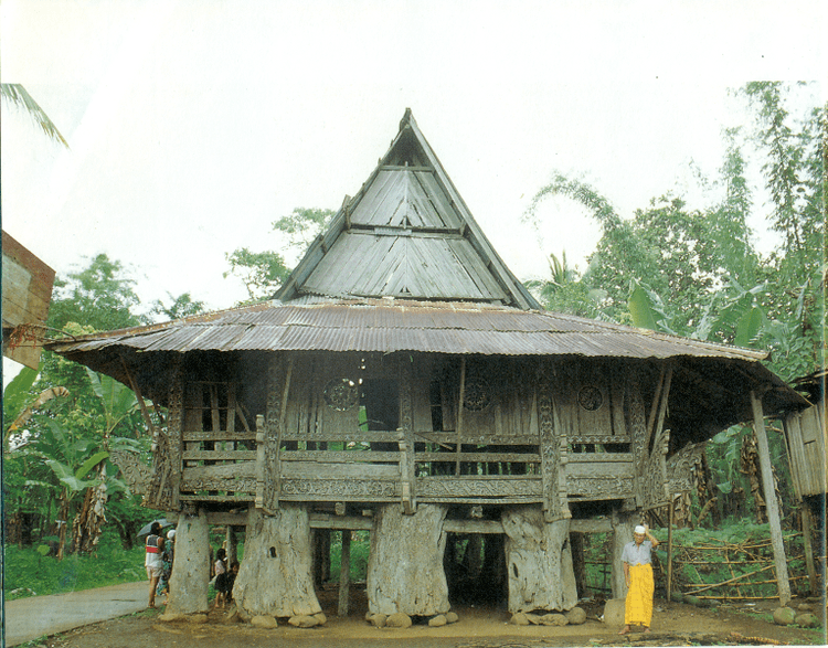 The Kawayan Torogan, a traditional Maranao torogan built by Sultan. There are group of people (left) under the Kawayan Torogan and a man (front, right). Kawayan Torogan is surrounded by trees with a basketball ring on the left side. The man (front, right) is wearing a white taqiyah, a gray polo, and a yellow cloth wrapped around his waist