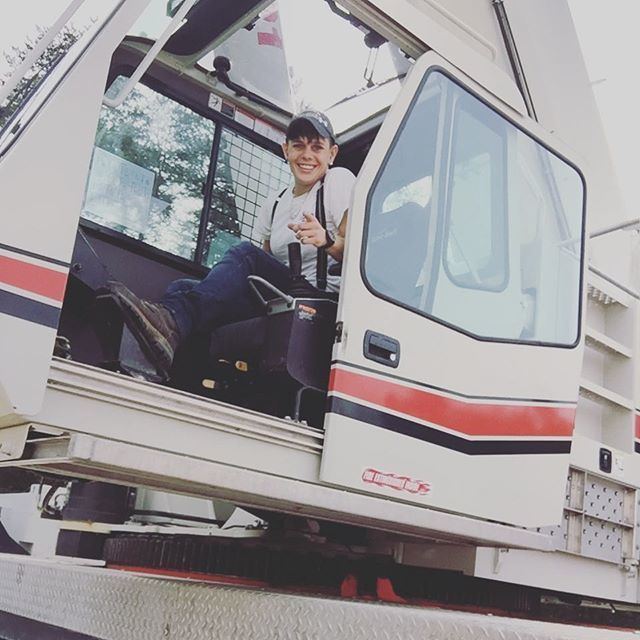 Jessica Middlebrook (Tow Truck Jess) smiling while inside a tow truck, wearing earrings, a black cap, a white shirt, blue jeans, and brown shoes.