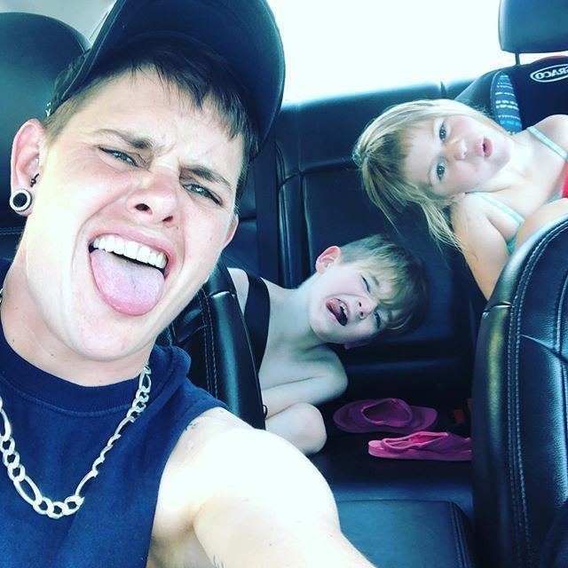 Jessica Middlebrook (Tow Truck Jess) doing a tongue-out gesture, wearing a black cap, earrings, a necklace, and sleeveless black shirt, while inside a car with her son Aryn and daughter Magie.