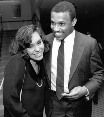 Tracie Ruiz smiling while wearing a long sleeve blouse while the man beside him wearing a coat, long sleeves, striped necktie, and pants