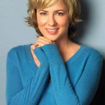 Traylor Howard smiling with short wavy blonde hair and clasping her hands while wearing a blue long sleeve knitted jacket