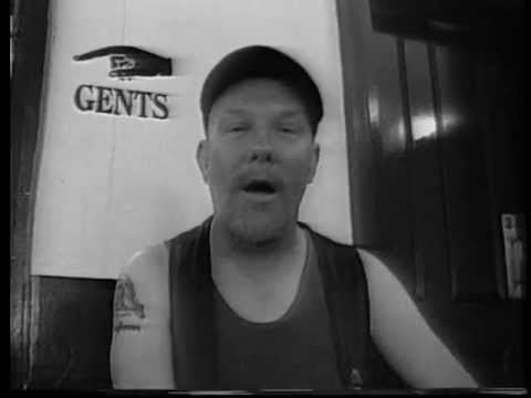 Troughman is serious, mouth half opened, has a beard, and a mustache, sitting down inside a room with a GENTS sign with a hand poking behind his right, and a door on his left, has a tattoo on his right arm, wearing a black cap, black sando under a sleeveless vest with a pin on left.