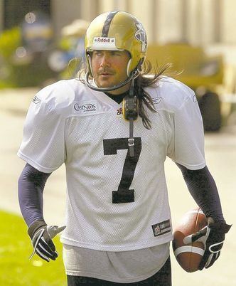 Troy Westwood with a serious face while holding a football, with long hair, wearing a white football uniform and a yellow protective hat.