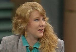A younger Truddi Chase with long hair speaking in an interview and wearing a gray blazer over a blue dress.