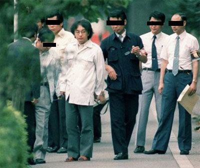 Tsutomu Miyazaki wearing eyeglasses, white long sleeves, green pants and escorted by policemen.