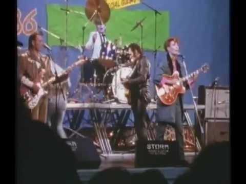 In the movie scene of Tutti Frutti 1987-Tv series, in a stage from left, Jake D’Arcy is serious standing playing his guitar has brown hair wearing a brown cowboy jacket and pants, 2nd from left, on the stage with drums Stuart McGugan is sitting, playing drums, 3rd from left, Maurice Roëves is standing, right side view, looking down playing his guitar, has black hair wearing a black leather jacket and a black pants, at the right, Emma Thompson is serious, singing, standing while playing her guitar, has orange hair wearing a black top with pink necktie under a blue coat and blue pants