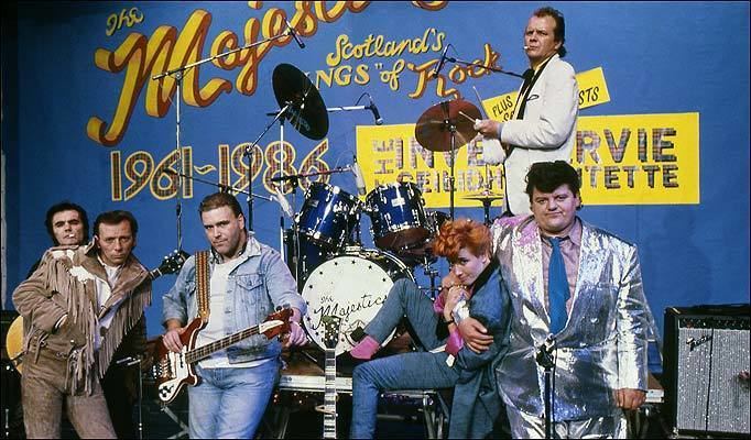 in the movie scene of Tutti Frutti (1987 TV series)in a studio with a blue wall with a drums at the back, from left, Maurice Roëves is serious with a cigarette in his mouth has black hair, 2nd from left Jake D’Arcy
is serious, standing, leaning to his left, with his hands on his pockets, and a guitar on his back, has black hair wearing a brown cowboy jacket and brown pants, 3rd from left, Ron Donachie is standing leaning backward, standing, holding his guitar, gas black hair wearing a white shirt under a denim jacket and denim pants, 4th from left, Emma Thompson is sitting, with his hands on the arm of Robbie, has orange hair, wearing a blue coat, black bracelet, blue pants, pink socks with black shoes, 5th from left, behind Emma, Stuart McGugan is serious with a cigarette in his mouth standing holding a drumstick has black hair wearing a white coat, at the right Robbie Coltrane is smiling, standing holding Emma Thompson with his right arm, has black hair wearing purple polo with blue necktie under a silver shiny coat and pants.