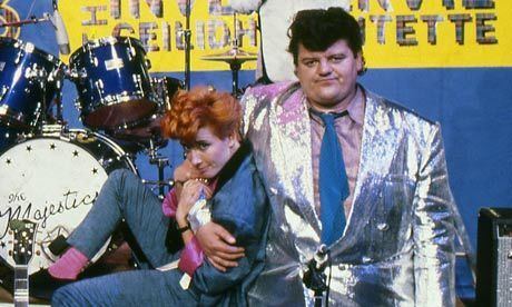 in the movie scene of Tutti Frutti (1987 TV series)in a studio with a blue wall with a drums at the back, from left, Emma Thompson is sitting, with his hands on the arm of Robbie, has orange hair, wearing a blue coat, black bracelet, blue pants, pink socks with black shoes, at the right Robbie Coltrane is smiling, standing holding Emma Thompson with his right arm, has black hair wearing purple polo with blue necktie under a silver shiny coat and pants.