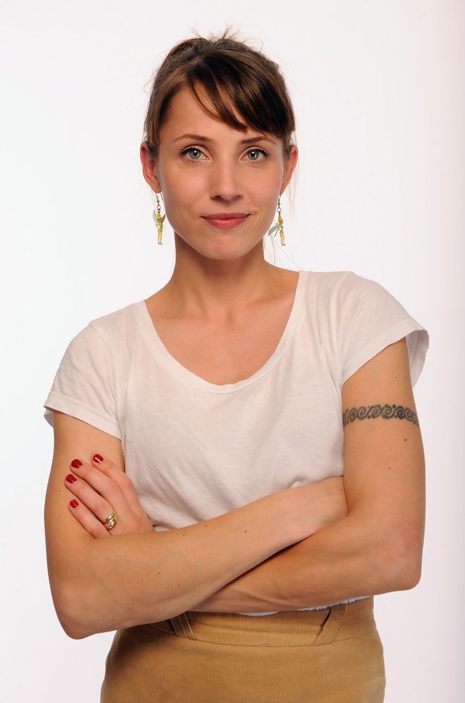 Tuva Novotny with a tight-lipped smile and her arms crossed showing her tattoo while wearing a white t-shirt, brown skirt, and earrings