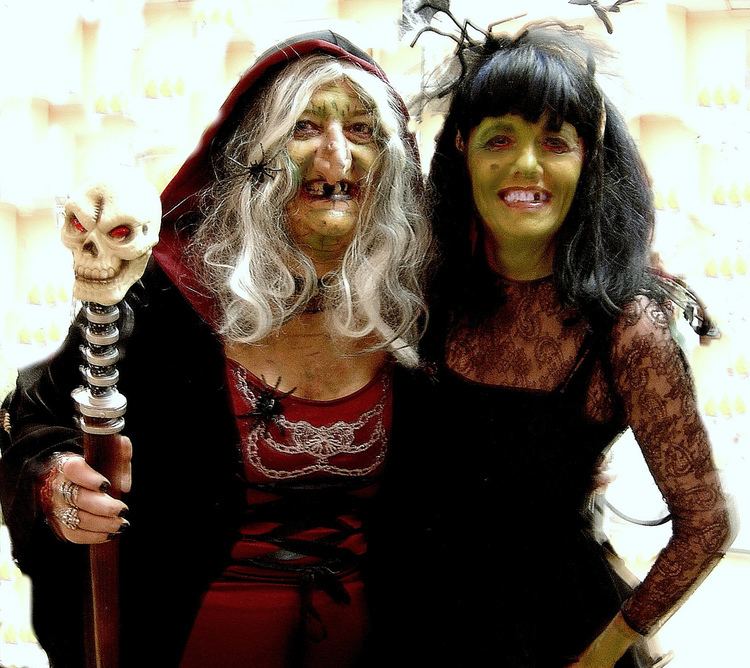 Wicca and Darlene Dranfield are smiling while wearing witch costumes. Wicca is holding a cane with a skull, with white wavy hair, and wearing a black and red cloak while Darlene is wearing headdress and black lace dress