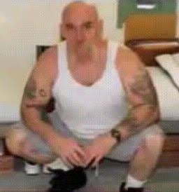 Tyler Bingham doing squat, with a bald head, with a tattoo on his body, wearing a sleeveless white shirt, gray shorts, and black shoes.