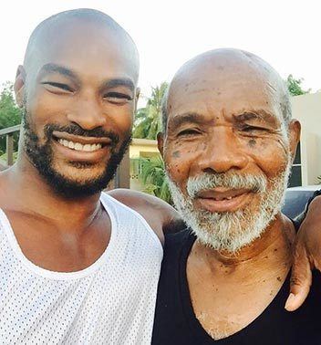 Tyson Beckford smiling with mustache and beard while wearing a white sando and his father wearing a black shirt
