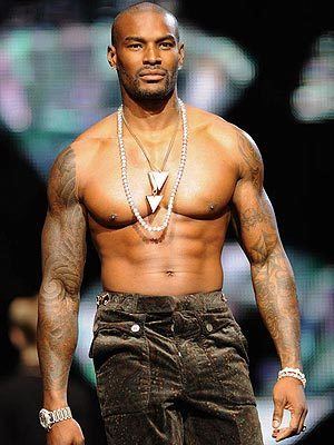 Tyson Beckford wearing brown pants, necklaces, a bracelet, and a wristwatch