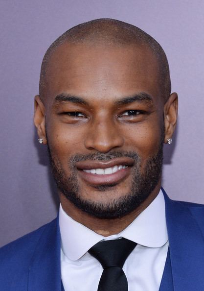 Tyson Beckford smiling with mustache and beard while wearing a blue coat, white long sleeves, black necktie, and earrings
