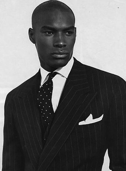 Tyson Beckford posing for Ralph Lauren Corporation while wearing a striped coat, white long sleeves, and polka dot necktie