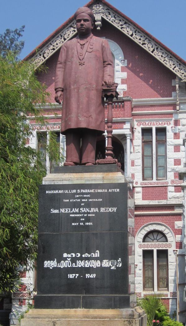 The statue of Ulloor S Parameswara Iyer in front of a maroon building, on the left is a bamboo tree leaves in the middle is Ulloor S Parameswara is serious, standing with his left hand on a table, wearing a turban, necklace and Sherwanis in maroon color statue, Below is a black marble pedestal with the words “Ulloor S Parameswara AIyer (1877-1949) This statue was unveiled by Shri. Neelam Sanjiva Reddy President of India on May 15, 1961.” below is the name of “Ulloor S Parameswara Iyer in Indian writing 1977-1949”
