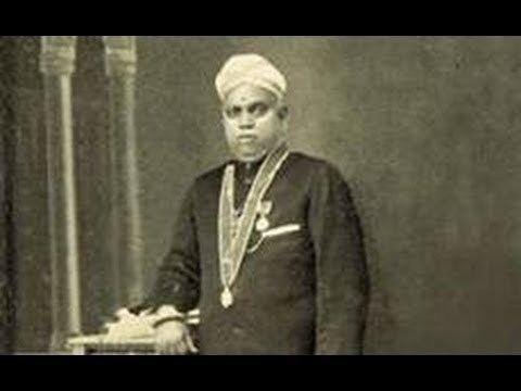 Ulloor S Parameswara is serious, standing with his right hand on the book on top of a table with two pillars at the back of his right arm, wearing a white turban, necklace, gold, silver medals and black Indian suit.