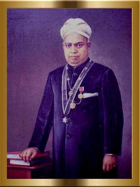 In a gold frame, Ulloor S Parameswara is serious, standing with his right hand on the book on top of a table, wearing a white turban, necklace, gold, silver medals and black indian suit.