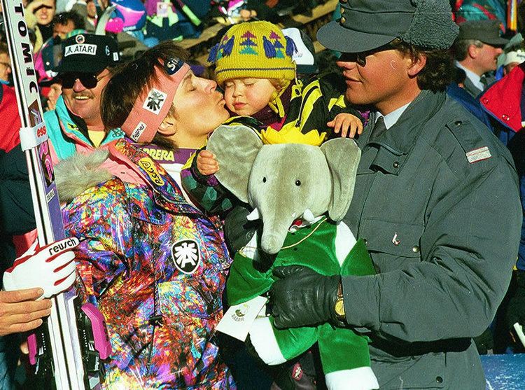 Ulrike Maier holding a snowboard and kissing her baby on the cheek with his husband while standing in the midst of the crowd. Ulrike is wearing a white shirt under a colored jacket, red and white gloves, and a headband. Her baby is wearing a black and yellow jacket and a yellow bonnet while her husband is carrying their baby and an elephant stuffed toy, wearing a black coat, black gloves, black hat, black sunglasses, and a watch.