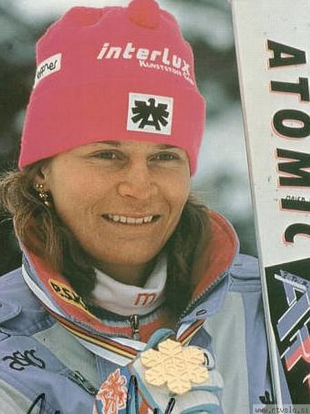 Ulrike Maier smiling while carrying her snowboard with the written word "ATOMIC", a turtleneck jacket with the written letter "m" on it under a blue and white coat, blue and white gloves, and a red bonnet hat with the printed word "interlux"