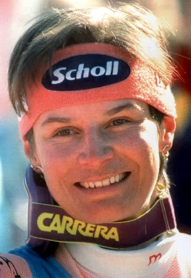 Ulrike Maier smiling with short hair and wearing a jacket with the printed letter "m" on the neck part, neck support with the printed word "CARRERA", headband with the printed word "Scholl" and ear piercing