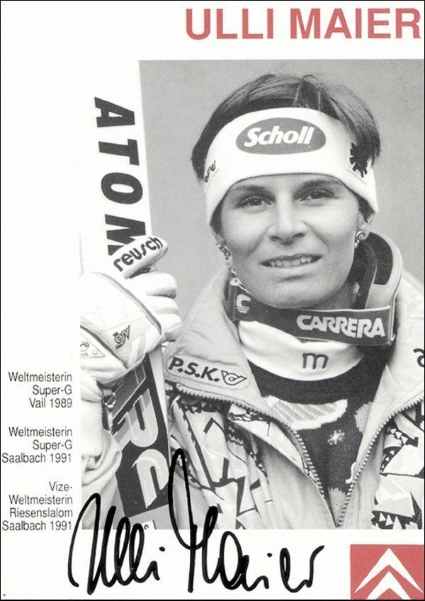 A poster of Ulrike Maier smiling and carrying a snowboard with the printed word "ATOMIC" and her name written at the top of the poster with her signature at the bottom, and the name of events she joined on the left side. Ulrike wearing a jacket with the letter "m" under a printed jacket, neck support with the printed word "CARRERA", a headband with the printed word "Scholl", gloves with the printed word "Reusch" and ear piercing