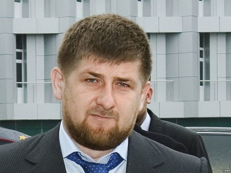 Umar Israilov looking serious with a beard and mustache and wearing a white shirt under a gray coat with a blue necktie