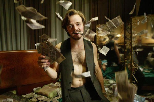 Matthew Newton smoking while holding money and wearing a black vest in a scene from the 2009 drama series, Underbelly: A Tale of Two Cities