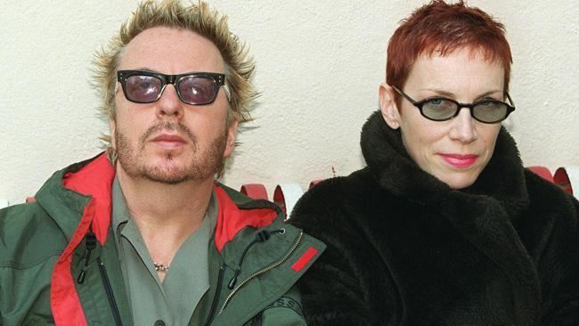 Annie Lennox smiling and wearing eyeglasses, a black scarf, and a black coat while Dave Stewart wearing a green and orange jacket