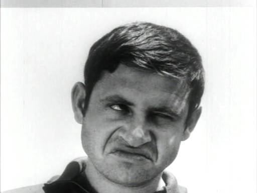 Uri Zohar doing a funny face, right eye looking from above, left eye is half closed, has black hair, beard, and mustache, wearing a black polo shirt.