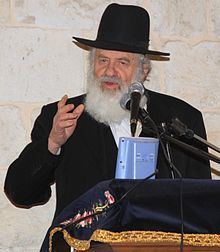 Uri Zohar, is seriously talking in a microphone, has white hair, a long white beard, and a mustache with right-hand pointing, left hand holding a blue Tanakh, in front of him is a lectern with a dark blue lectern cloth that has an embroidered design of a pink flower and yellow strips along the edge, he is wearing white long sleeves under a black frock coat and a black shtreimel.