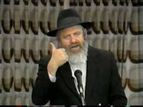 Uri Zohar is seriously talking in a microphone, has white hair, a white beard, and a mustache, his right hand doing the thumbs-up, has a microphone in front, wearing a black shtreimel, white long sleeves under a black frock coat.