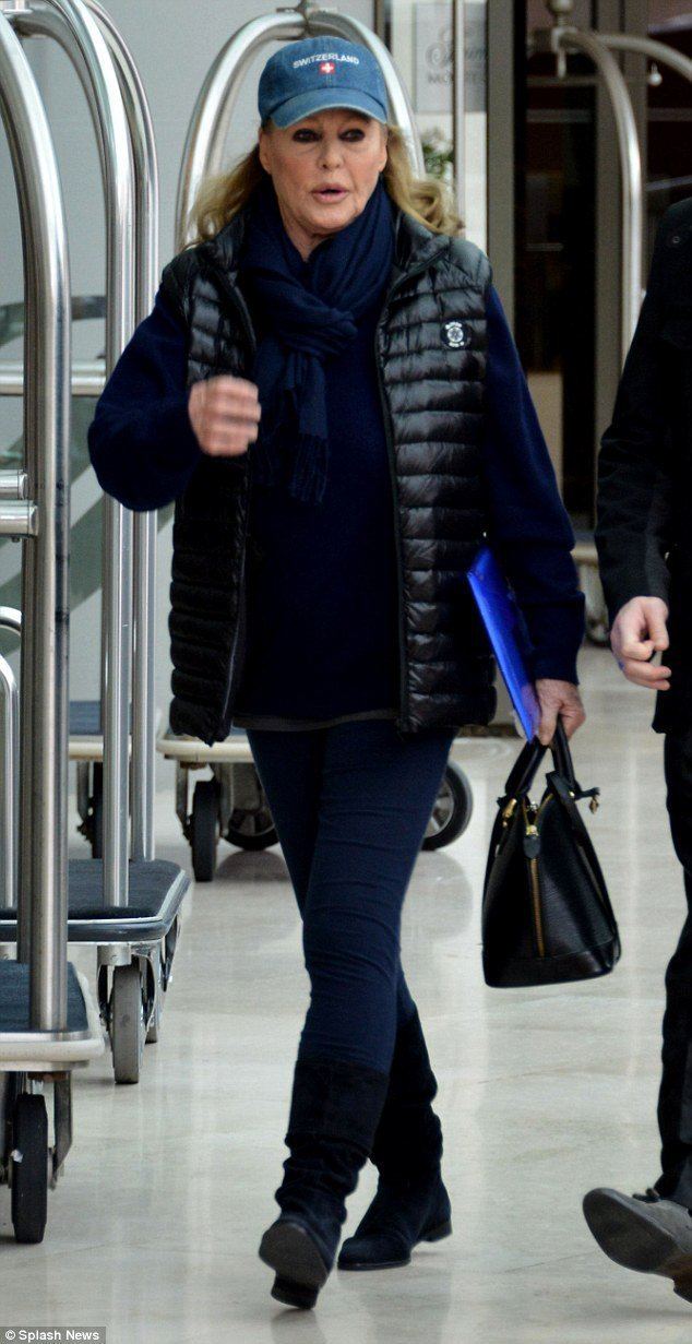 Ursula Andress in an airport wearing a blue cap and a black jacket over blue clothes and holding a bag and plastic envelope.