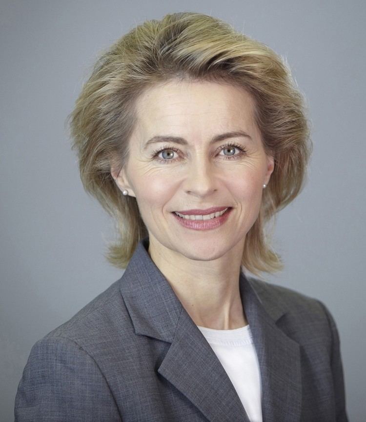 Ursula von der Leyen smiling, with short blonde hair, wearing pearl earrings and a gray blazer over a white top.