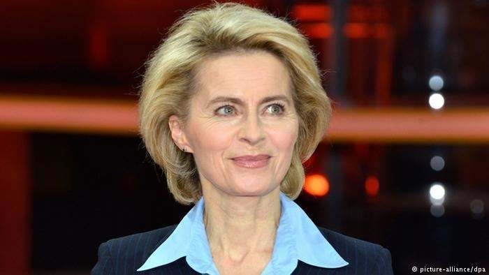 Ursula von der Leyen with a tight-lipped smile, short blonde hair, wearing a dark blue blazer over a light blue polo shirt.