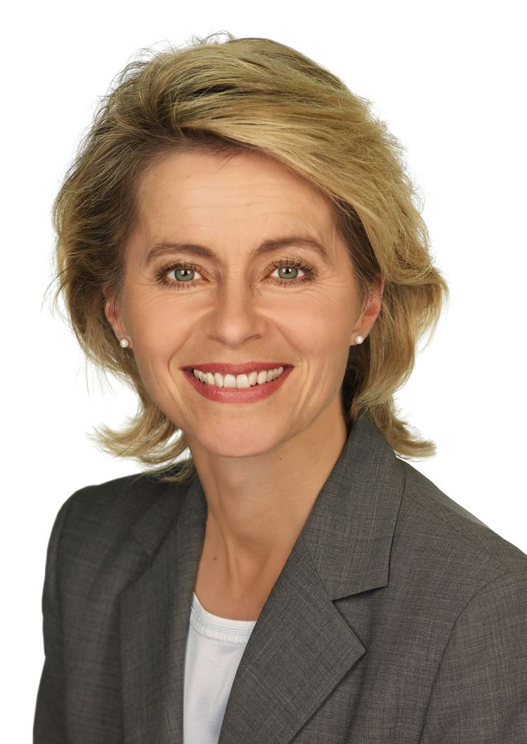 Ursula von der Leyen smiling, with short blonde hair, wearing pearl earrings and a gray blazer over a white top.