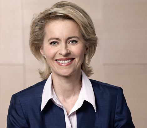 Ursula von der Leyen smiling, with short blonde hair, wearing earrings, a dark blue blazer over a white polo shirt.