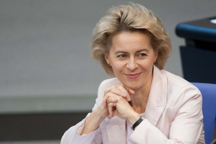 Ursula von der Leyen with a tight-lipped smile while holding her hands together and looking at something, with short blonde hair, wearing a black watch, and an off-white blazer over white top.