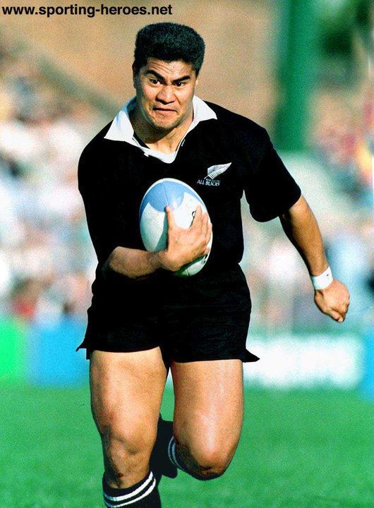 Va'aiga Tuigamala running while holding a rugby ball and wearing a black and white shirt, white wrist band, black shorts, and black socks with white stripes