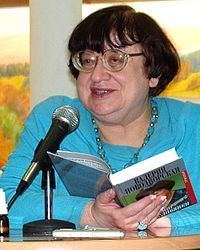 Valeriya Novodvorskaya speaking through a microphone while holding a history book, she has a short black hair with bangs, wearing an eyeglass, blue beads necklace, a blue ring on her right ring finger, and a blue long sleeve